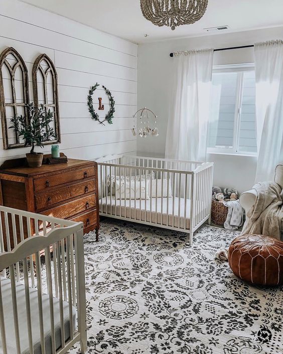 shared farmhouse nursery with a printed rug, a leather ottoman, a stained dresser, a bead chandelier and some greenery