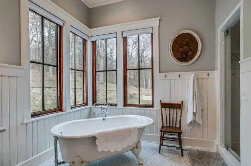 Corner bathroom features a freestanding clawfoot tub angled in front of a bank of French windows, a wooden slat back chair, hex tile flooring, and beadboard walls.