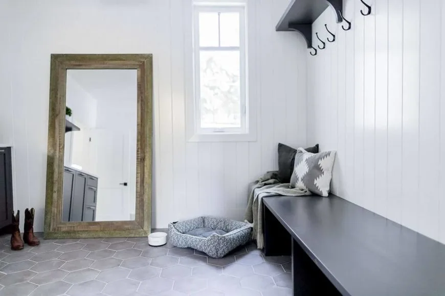 Farmhouse-style mudroom features a long wooden bench with space underneath for storage and black shelving with hooks overhead that stand out against the white plank wall. The space also includes a full-length mirror resting on the marble hexagon tile floor